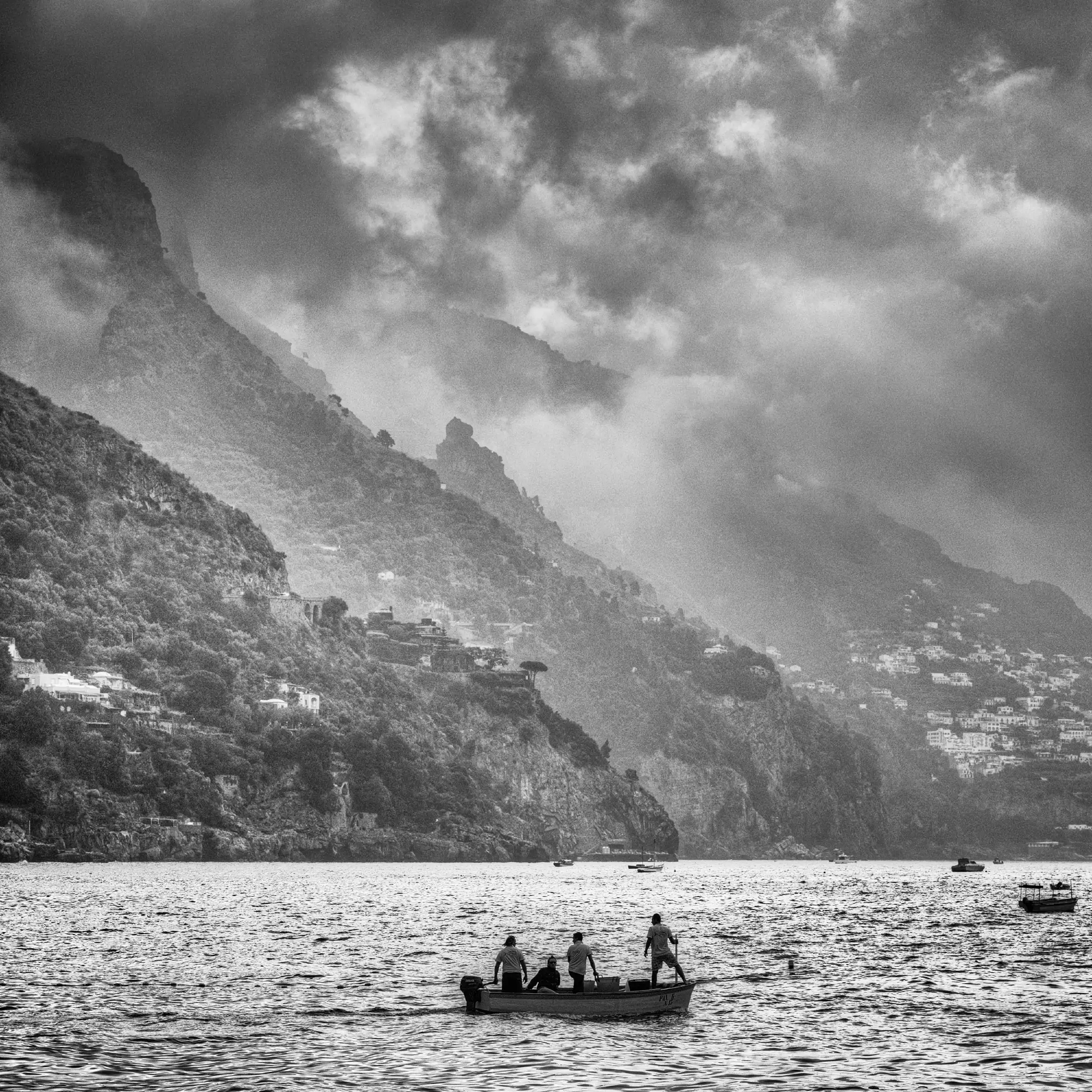 Black and white photograph of fishing boat with four crew members on calm Mediterranean waters, with dramatic cloudy sky above and terraced hillside village of Positano, Italy in background.