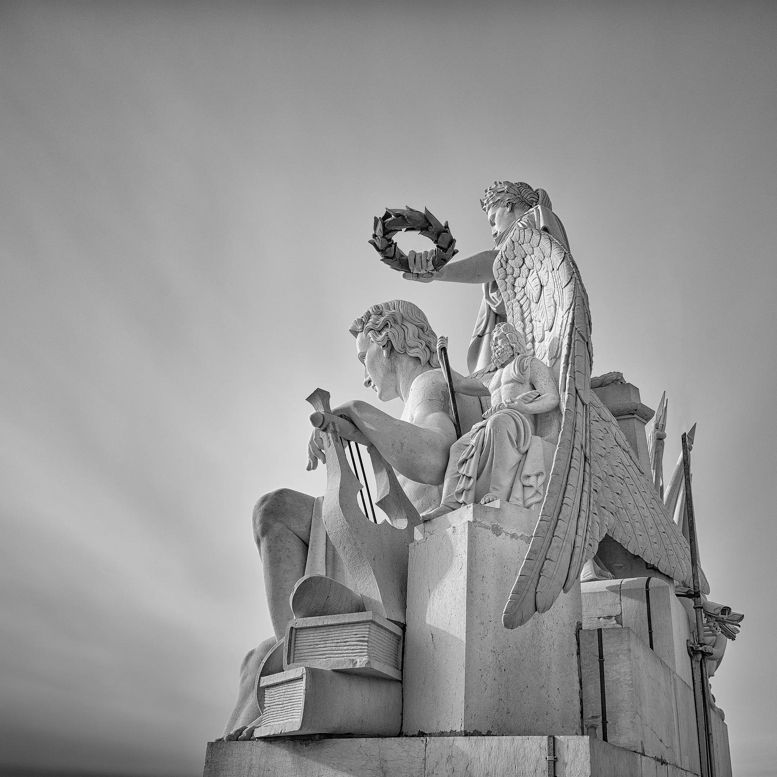 Black and white photograph of classical stone sculptures atop the Rua Augusta Arch in Lisbon, featuring draped figures with laurel wreaths against a dramatic sky