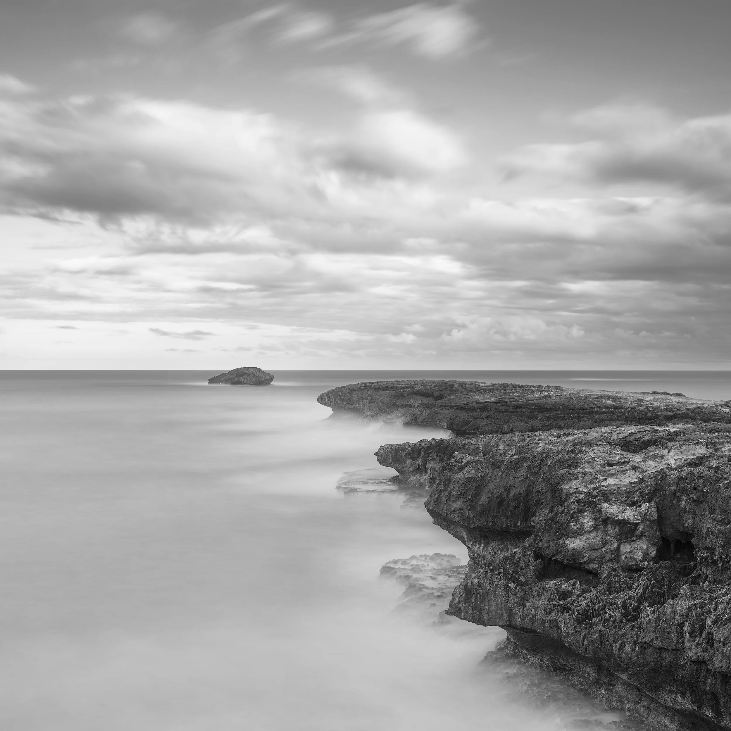 Black and white long exposure photograph of rocky volcanic coastline at Lāʻie Point, Oahu, showing dramatic rock formations extending into misty ocean waters under a cloudy sky.