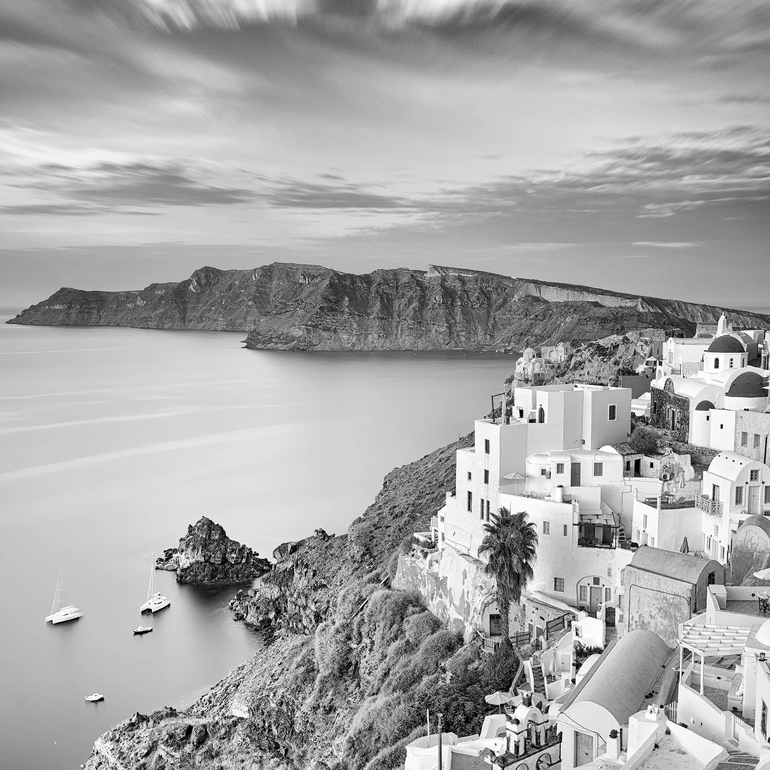 Black and white photograph of traditional Greek architecture cascading down clifftops in Oia, Santorini, with dramatic sky and boats in the harbor below